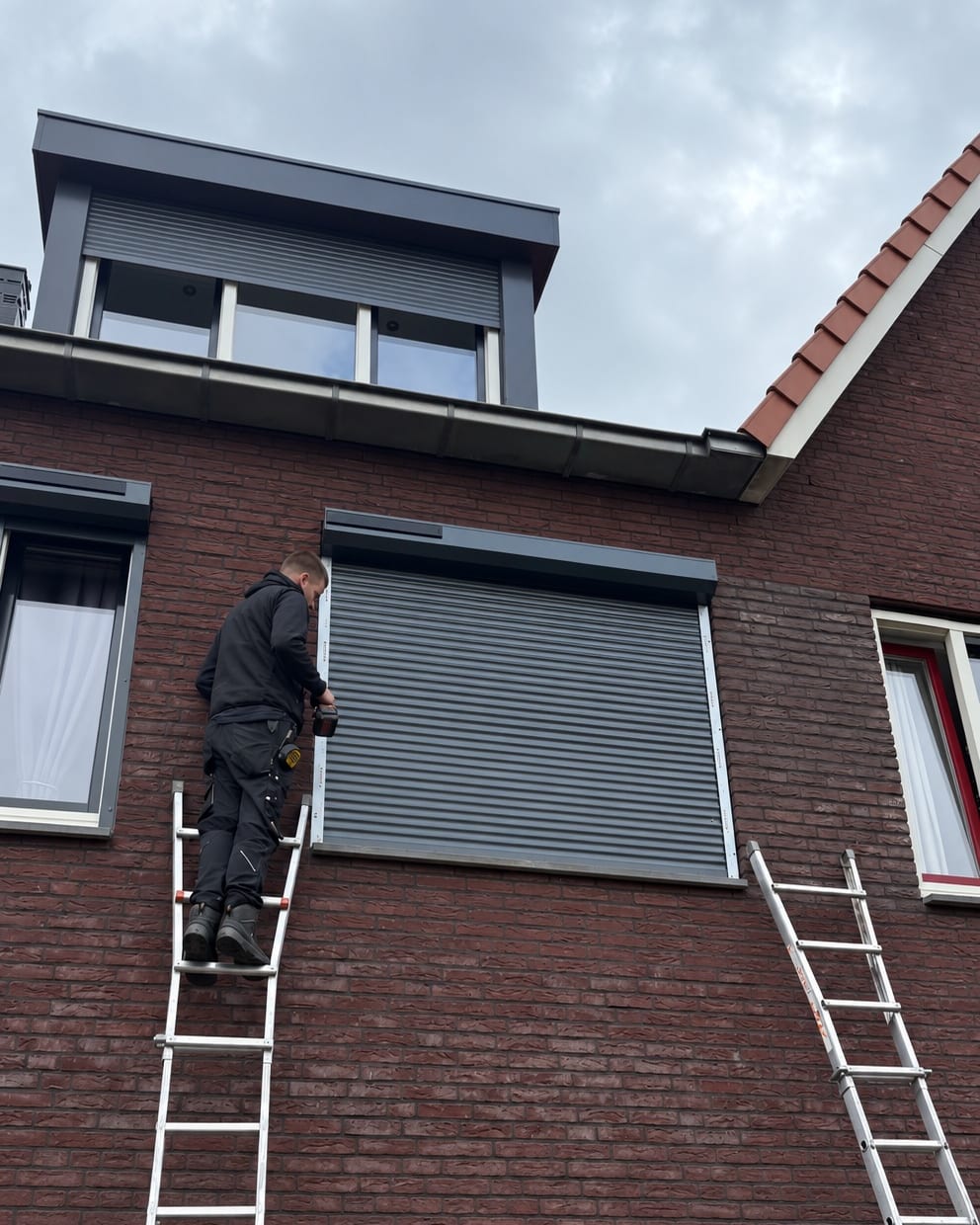 Bas staat op een ladder en schroeft een rolluik vast aan de voorgevel